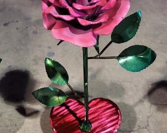 May include: A pink metal rose with green leaves stands on a red metal heart. The rose and heart are both shiny and have a metallic finish.