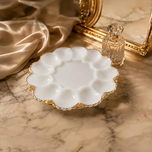 Pode incluir: Um prato para ovos de vidro leitoso branco com borda recortada e acabamento dourado. O prato possui doze cavidades circulares dispostas em anel ao redor de um centro liso. Um pequeno frasco de perfume de vidro facetado com gargalo dourado.