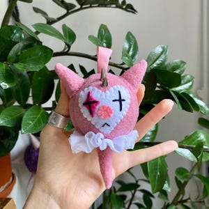 May include: A pink felt ornament in the shape of a cartoon character with a heart-shaped face and a white ruffled collar. The character has a sad expression, a pink nose, and a black cross and star design on its face. Silver spikes adorn the head.