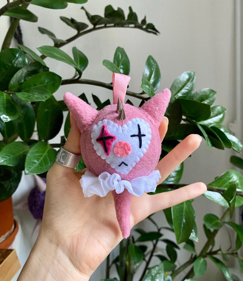 May include: A pink felt ornament in the shape of a cartoon character with a heart-shaped face and a white ruffled collar. The character has a sad expression, a pink nose, and a black cross and star design on its face. Silver spikes adorn the head.