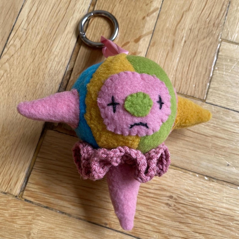 May include: A colourful felt keychain in the shape of a clown. The character has a pink face with black cross eyes and a sad mouth, a green nose, and a ruffled pink collar. The body is a patchwork of yellow, green, blue, and pink. Includes a silver keyring.