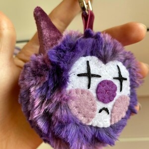 May include: A plush keychain featuring a purple, furry creature with a sad clown face. The face has white, pink, and purple felt details, including cross-shaped eyes and a circular nose. A purple cone and a silver clasp complete the design.