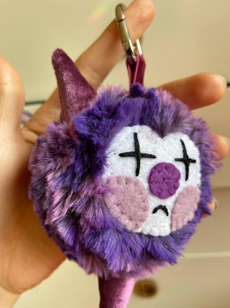 May include: A plush keychain featuring a purple, furry creature with a sad clown face. The face has white, pink, and purple felt details, including cross-shaped eyes and a circular nose. A purple cone and a silver clasp complete the design.