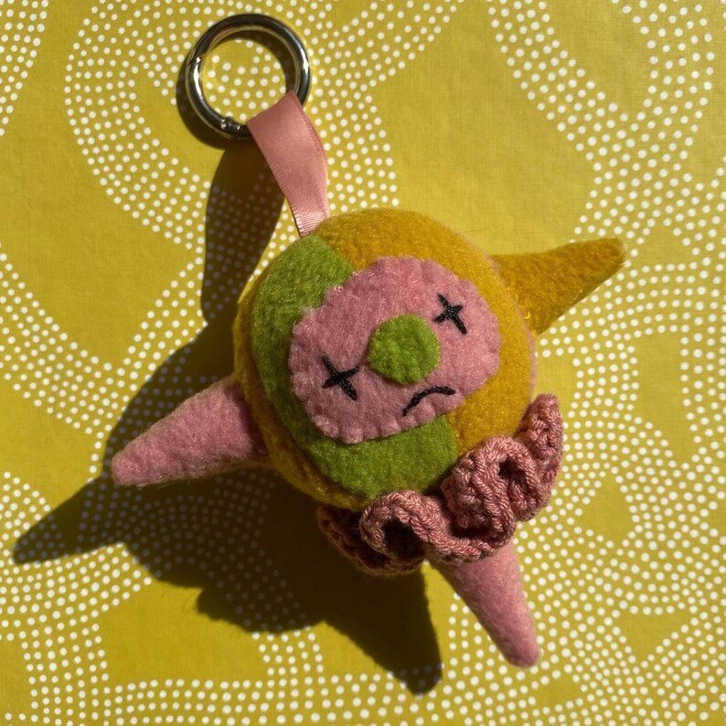 May include: A handmade felt clown keyring with a metal ring and pink ribbon. The clown has a yellow and green head, pink face and nose, black cross eyes, and a pink ruffled collar. The clown's arms and nose are pink.