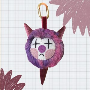May include: A quirky plush keychain in the shape of a monster's face. The face is a mix of purple and pink fur, with white and pink facial features. It has a gold-coloured clasp and a burgundy velvet strap. The keychain is designed with a sad expression.