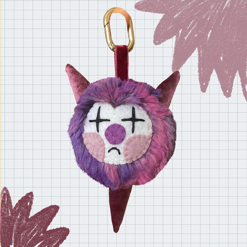 May include: A quirky plush keychain in the shape of a monster's face. The face is a mix of purple and pink fur, with white and pink facial features. It has a gold-coloured clasp and a burgundy velvet strap. The keychain is designed with a sad expression.
