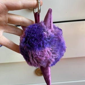 May include: A purple and pink plush keychain in the shape of a stylized creature. The keychain has a fluffy, round body with two pointed ears and a long, pointed tail. It is attached to a silver clasp with a purple strap.