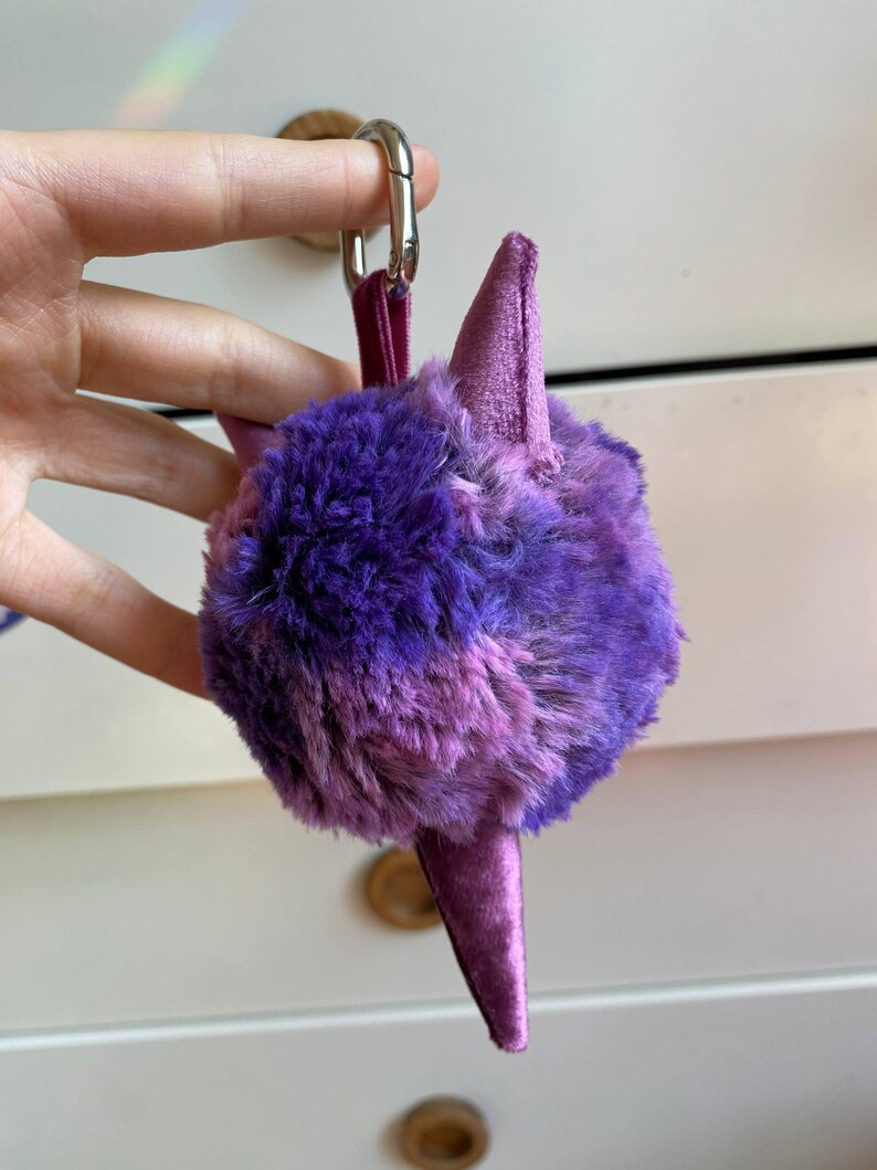 May include: A purple and pink plush keychain in the shape of a stylized creature. The keychain has a fluffy, round body with two pointed ears and a long, pointed tail. It is attached to a silver clasp with a purple strap.