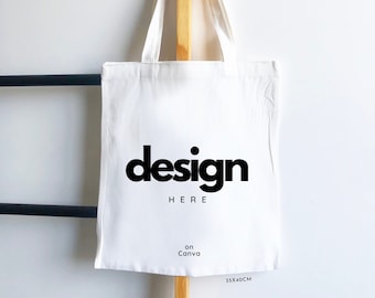 Puede incluir: Una bolsa de tela blanca con dos asas, que presenta el texto impreso en negro "design HERE" en el centro y "on Canva" debajo. La bolsa incluye una pequeña etiqueta en la esquina inferior derecha con el texto "35X40CM" indicando su tamaño.