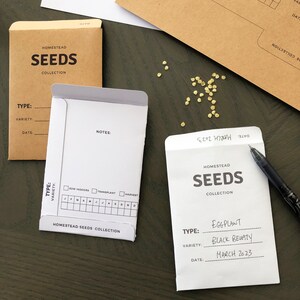 May include: Two white seed packets with black text. The top packet says "Homestead Seeds Collection" and the bottom packet says "Homestead Seeds Collection" with the text "Type: Eggplant, Variety: Black Beauty, Date: March 2023".