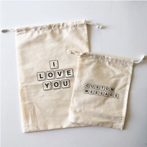 Könnte beinhalten: Zwei beigefarbene Kordelzugbeutel. Der größere Beutel hat die Worte "I LOVE YOU" mit schwarzen Buchstabenkacheln geschrieben. Der kleinere Beutel hat die Worte "CUSTOM MESSAGE" mit schwarzen Buchstabenkacheln geschrieben.