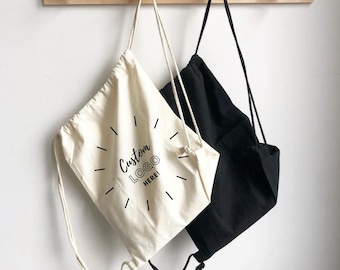 May include: Two drawstring backpacks, one cream and one black, featuring thin rope straps. The cream bag displays the black text "Custom LOGO HERE!" surrounded by a circular sunburst graphic. Both bags are suspended from a wooden wall hook.