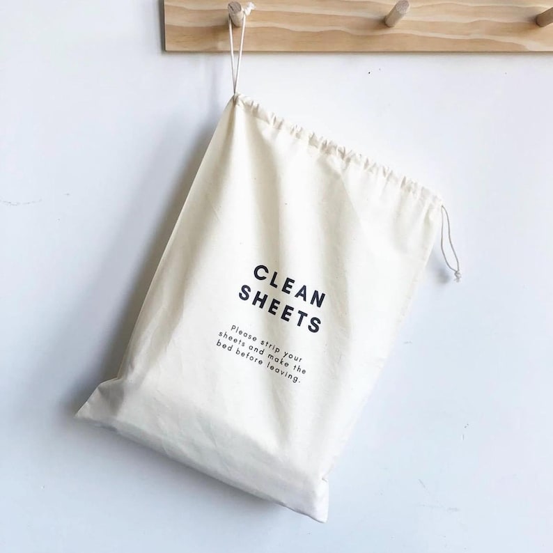May include: A cream-colored fabric drawstring bag featuring the black text CLEAN SHEETS followed by the smaller text Please strip your sheets and make the bed before leaving. The bag is suspended by its drawstring cord from a wooden wall hook.