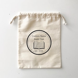 May include: A beige drawstring bag with a black circular logo. The logo features the text "Custom Name Book Club" around a dotted circle, with an open book illustration and the year "est. 2025" below.