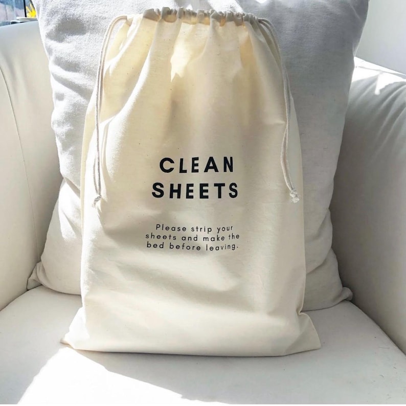 May include: An off-white drawstring laundry bag made of fabric. The bag features the black text CLEAN SHEETS in a bold sans-serif font, followed by the smaller black text: Please strip your sheets and make the bed before leaving.