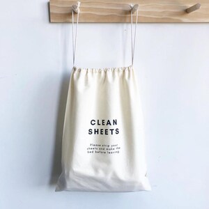 May include: A cream-colored cotton drawstring laundry bag featuring the black text CLEAN SHEETS followed by the smaller printed instruction: Please strip your sheets and make the bed before leaving. The bag has two long white cords for hanging.