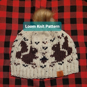 Op de afbeelding: Een witte en bruine gebreide muts met een pompon bovenop. De muts heeft een bruin en zwart patroon van bomen. De muts ligt op een rood-zwarte geruite achtergrond. De tekst "Loom Knit Pattern" staat boven de muts. De muts heeft een bruin lederen label met de tekst "Local Made".