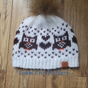 May include: White knitted hat with a brown and orange owl pattern and a brown faux fur pom pom. The hat has a brown leather tag with the text "Loom Knit Pattern".