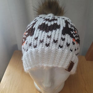 May include: White knit beanie with a brown and white geometric pattern and a brown faux fur pom pom on top. The beanie has a brown leather strap on the side.
