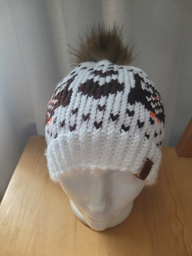 May include: White knit beanie with a brown and white geometric pattern and a brown faux fur pom pom on top. The beanie has a brown leather strap on the side.