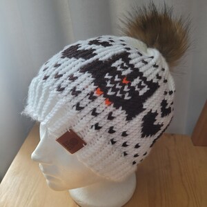 May include: White knit beanie with a brown and orange geometric pattern and a brown faux fur pom pom. The beanie has a brown leather tag with the word "Made" on it.