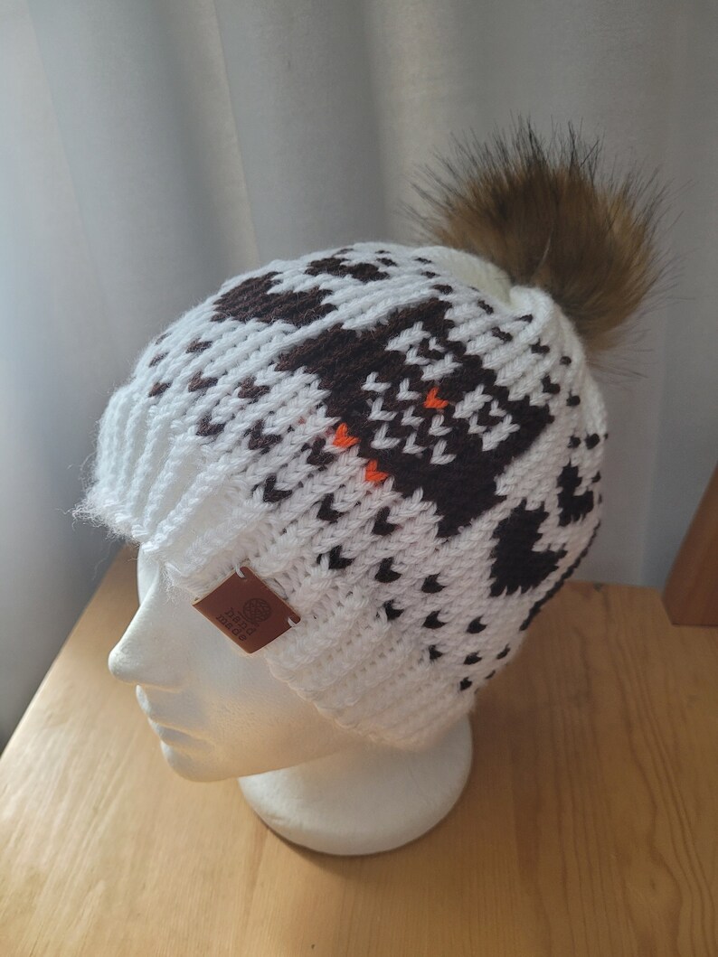 May include: White knit beanie with a brown and orange geometric pattern and a brown faux fur pom pom. The beanie has a brown leather tag with the word "Made" on it.