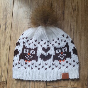 May include: White knitted beanie with a brown faux fur pom pom and a brown owl pattern. The beanie has a brown leather tag with the word "Owl" on it.