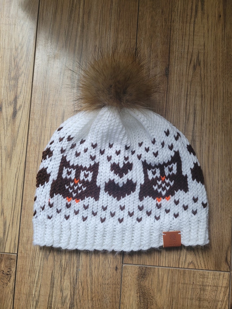 May include: White knitted beanie with a brown faux fur pom pom and a brown owl pattern. The beanie has a brown leather tag with the word "Owl" on it.