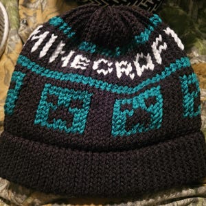May include: A dark navy blue knit beanie featuring the word "MINECRAFT" in white and teal. The hat has a folded brim and a repeating pattern of teal and black pixelated squares. The beanie is designed for warmth and style.