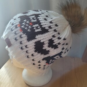 May include: White knit beanie with a black and brown geometric pattern and a brown faux fur pom pom.