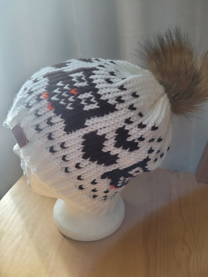 May include: White knit beanie with a black and brown geometric pattern and a brown faux fur pom pom.