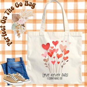 May include: Cream tote bag featuring a watercolour design of red and pink hearts on stems. The text "Love never fails" and "I Corinthians 13:8" are printed below the hearts. The bag is displayed with jeans and trainers.