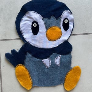 May include: A fabric appliqué shaped like a penguin, constructed from dark blue denim and light blue fabric pieces. It features a bright yellow felt beak and feet, two white button eyes with black fabric pupils, and visible zigzag stitching along the edges.