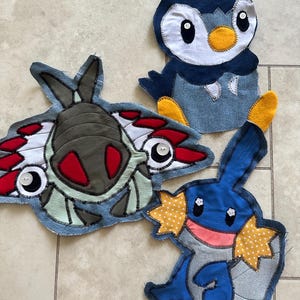 May include: Three fabric appliqués featuring cartoon creatures. One is a dark blue and white penguin-like figure with yellow feet. Another is a grey, red, and white insect-like creature. The third is a blue creature with yellow polka-dot fins.
