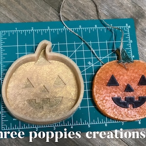 May include: A close-up of two Halloween-themed pumpkin decorations. One is a beige plastic mold with a jack-o'-lantern face, and the other is a round orange pumpkin with a black jack-o'-lantern face. The pumpkins are on a green cutting mat with a ruler.
