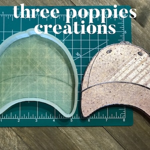 May include: A clear silicone mold of a baseball cap, next to a finished resin baseball cap with a brown and white striped pattern. The mold is on a green cutting mat with inch markings. The finished cap is on a wooden surface. The text "three poppies creations" is visible in the image.