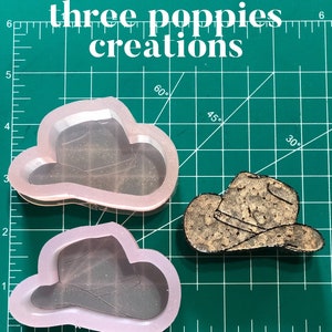 May include: Two silicone molds for making cowboy hats. The molds are pink and translucent. The molds are on a green cutting mat with white markings.