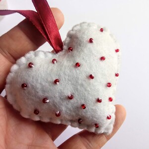 May include: White felt heart-shaped ornament with red beads sewn on. The ornament has a red ribbon attached to the top.