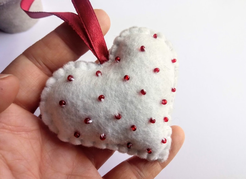 May include: White felt heart-shaped ornament with red beads sewn on. The ornament has a red ribbon attached to the top.
