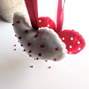 May include: Two felt hearts, one white and one red, are sewn together and decorated with red beads. The hearts are hanging from a red ribbon.