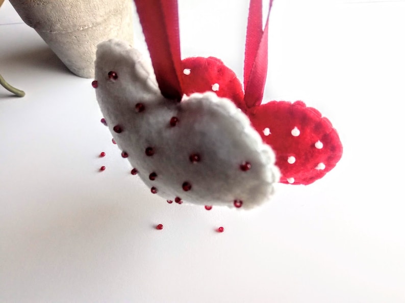 May include: Two felt hearts, one white and one red, are sewn together and decorated with red beads. The hearts are hanging from a red ribbon.