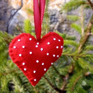 May include: A red felt heart with white polka dots hanging from a red ribbon. The heart is hanging from a green pine tree branch.