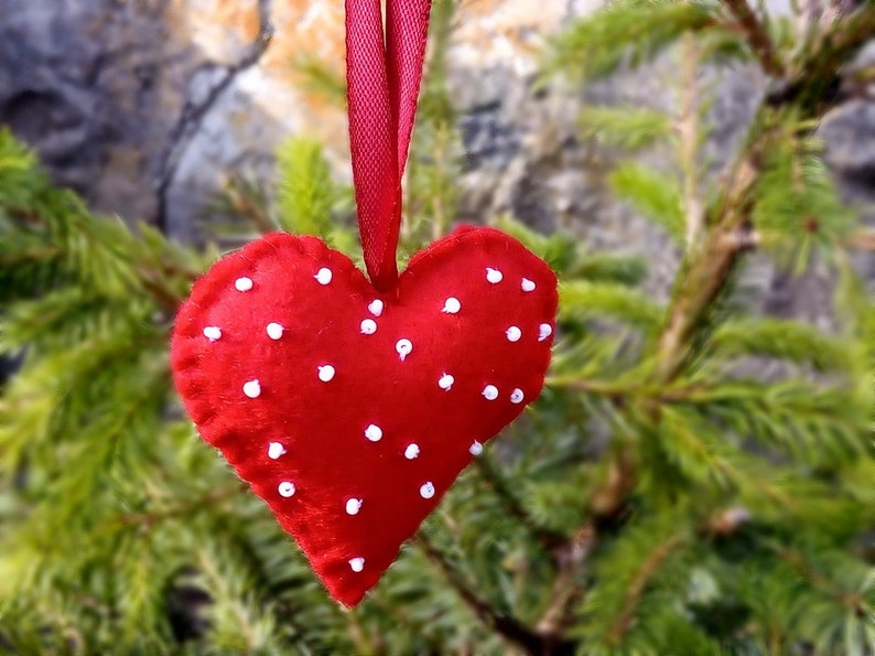 May include: A red felt heart with white polka dots hanging from a red ribbon. The heart is hanging from a green pine tree branch.