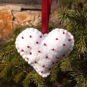 May include: White felt heart ornament with red beads sewn on. The ornament is hanging from a red ribbon on a green pine tree branch.