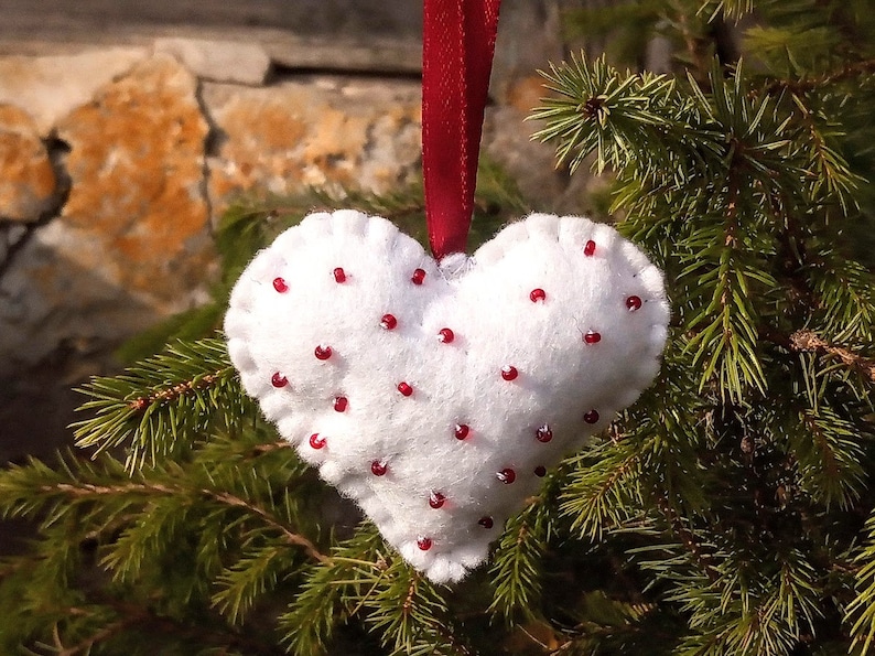May include: White felt heart ornament with red beads sewn on. The ornament is hanging from a red ribbon on a green pine tree branch.