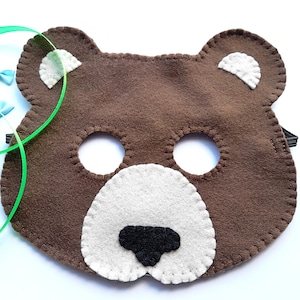 May include: A brown felt bear mask with white details on the ears and muzzle, and a black nose. The mask features eye holes and an elastic strap. Green ribbon and small blue bows are also visible.