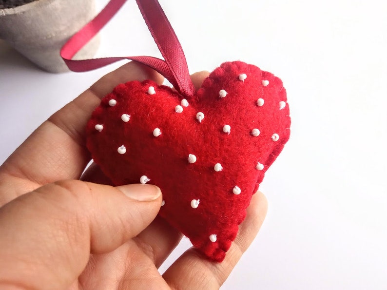 May include: A red felt heart with white polka dots, hanging from a red ribbon. The heart is handmade and has a soft, plush texture.