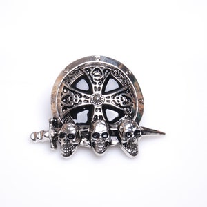 Puede incluir: Broche plateado con un diseño de escudo, una cruz ornamentada, tres calaveras y una espada. El broche tiene un tema gótico o medieval, con detalles intrincados y un centro oscuro contrastante.