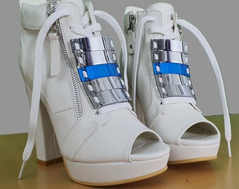 May include: A pair of white platform shoes with open toes and silver accents. The shoes have laces and a zipper on the side.