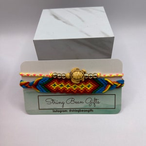 May include: Two handmade friendship bracelets. One bracelet is a colorful woven design with a turtle charm and silver beads. The other bracelet is a neon yellow, pink, and blue braided design. The bracelets are displayed on a card that reads "String Bean Gifts".
